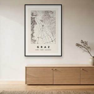 May include: A framed topographic map print of Graz, Austria, featuring a black and white detailed street grid and terrain elevation lines. The print includes the text GRAZ, coordinates 47.0708 N 15.4385 E, and contour interval 5 m in black.