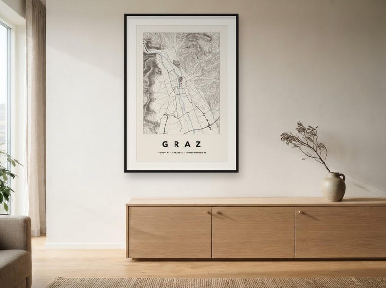 May include: A framed topographic map print of Graz, Austria, featuring a black and white detailed street grid and terrain elevation lines. The print includes the text GRAZ, coordinates 47.0708 N 15.4385 E, and contour interval 5 m in black.