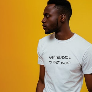 May include: A person wearing a white t-shirt with the text "GEH BUDDL DI NET AUF!" printed on it. The person is standing in front of a yellow background.