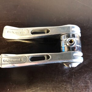 Op de afbeelding: Een zilverkleurige Shimano fiets-buisverstellerset met twee metalen hendels met langwerpige uitsparingen. Het centrale bevestigingsmechanisme bevat een klein rond boutgat en een gepolijste metalen afwerking op de behuizing.