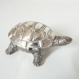 Pode incluir: Estatueta de tartaruga prateada com um padrão de concha detalhado e pernas e cabeça texturizadas. A estatueta é feita de metal e tem um acabamento brilhante. A tartaruga é um item decorativo.