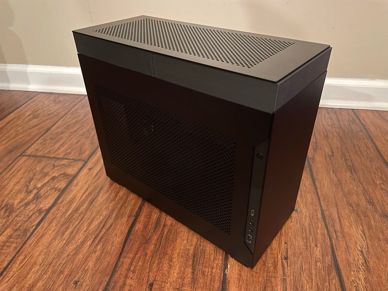 May include: A black computer case with a mesh front panel and a top panel with a mesh design. The case has a USB port and a headphone jack on the front.