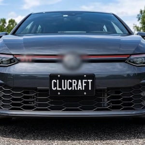 Puede incluir: Vista frontal de un coche gris oscuro con una matrícula negra que muestra el texto blanco CLUCRAFT. El vehículo presenta una rejilla inferior con patrón de panal, faros LED afilados y una fina línea roja horizontal en la parrilla delantera.