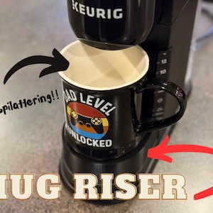 May include: A black coffee maker with a mug riser attachment. The mug riser is a black plastic platform that raises the mug up to the coffee spout. A black mug with a white rim and a graphic of a video game controller with the text "Dad Level Unlocked" is on the riser. The text "Less splattering!" is written in black on a white arrow pointing to the mug.