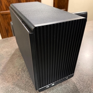 Puede incluir: Una caja de ordenador negra con una parte superior texturizada y un panel frontal con ranuras verticales. La caja tiene forma rectangular y parece estar hecha de metal. Varios puertos USB son visibles en la parte inferior del panel frontal.