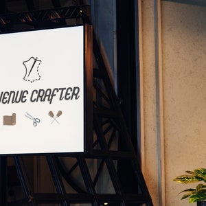 May include: A white sign with black text that reads "Avenue Crafter". The sign has a logo of a piece of leather with a needle and thread, a roll of leather, scissors, and two wooden tools.