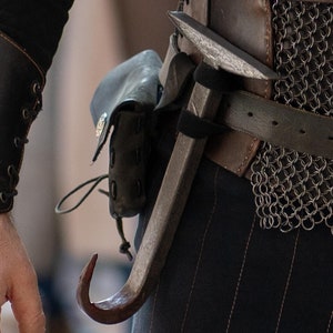 Może przedstawiać: Osoba ubrana w czarną koszulę i spodnie z brązowym skórzanym paskiem i spódnicą z kolczugi. Do paska przymocowany jest metalowy hak.