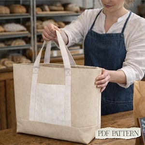May include: A large, rectangular tote bag made of light beige fabric, featuring two sturdy woven handles and a central exterior pocket with a subtle white vertical pinstripe pattern. The bag has a structured base and clean, simple lines.