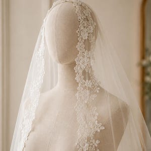Puede incluir: Un velo de novia blanco y transparente con un intrincado borde de encaje floral. El delicado patrón de encaje consiste en pequeñas flores y hojas, creando un borde festoneado que cae sobre la cabeza y los hombros de un maniquí.