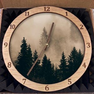 May include: A round wall clock with a light-colored wooden frame and a printed face featuring a dark green forest of pine trees against a foggy, muted background. The clock has dark brown hour and minute hands and engraved black numbers.
