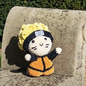 May include: A handmade crochet doll of a character with yellow hair, a black headband, and an orange and black outfit. The doll is sitting on a stone bench. The doll's face has black markings and a smiling expression.
