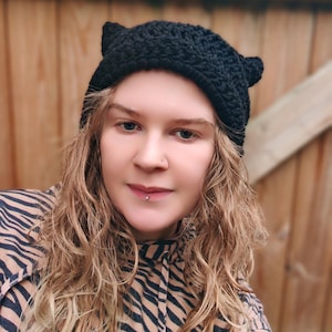 May include: A black crochet hat with cat ears. The hat is made of a textured yarn and is worn by a person with long, wavy blonde hair. The person is wearing a zebra-striped top.