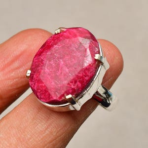 May include: A large, oval-cut ruby gemstone ring. The ruby is a vibrant, deep pink-red color, set in a silver-colored metal band. The ring's design features a simple, elegant setting that highlights the gemstone's size and brilliance.