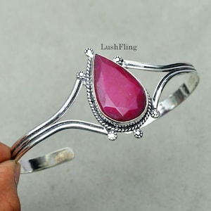 May include: A silver cuff bracelet featuring a central teardrop-shaped faceted dark pink gemstone. The metal setting has a twisted rope border and decorative silver beads. The text LushFling is printed above the gemstone on the bracelet.