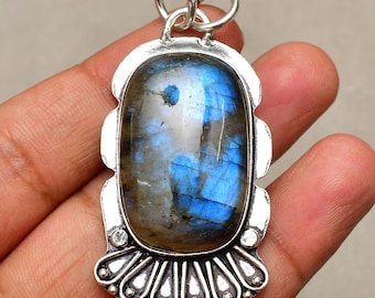 Puede incluir: Colgante de plata con una piedra labradorita ovalada. La piedra muestra tonos azules y grises iridiscentes. El colgante tiene un marco de plata decorativo y un lazo para una cadena. Diseño ornamentado y llamativo.
