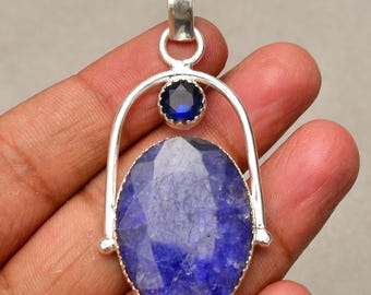 Op de afbeelding: Een zilveren hanger met een grote, ovaalvormige, gefacetteerde blauwe edelsteen onder een kleinere, ronde, gefacetteerde blauwe edelsteen. De stenen zitten in een zilveren boogframe dat verbonden is met een zilveren oogje aan de bovenkant.