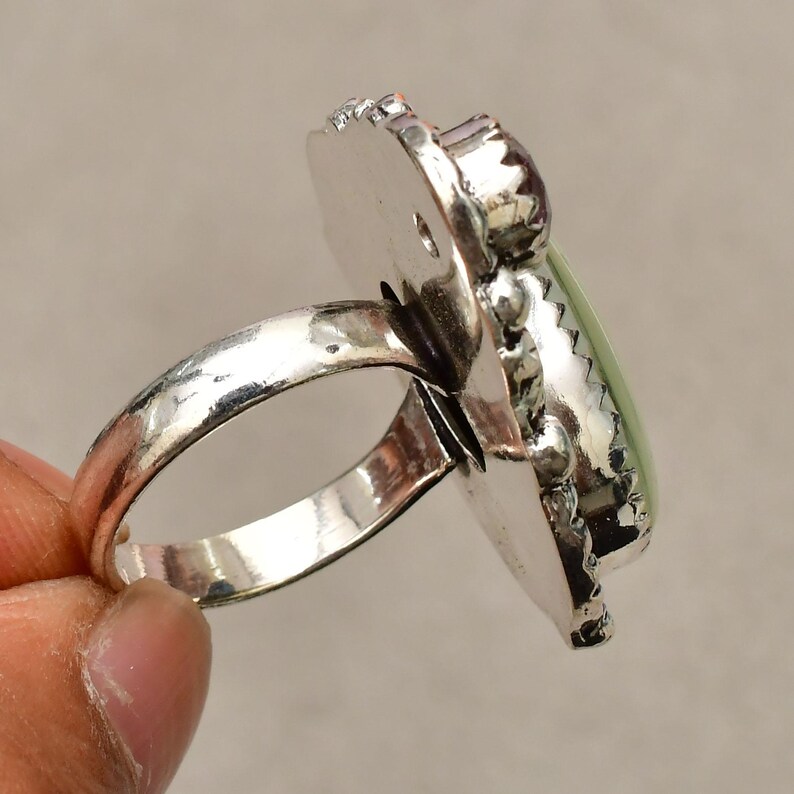 May include: A silver ring featuring a large, oval, pale green gemstone. The ring has a textured band and a decorative setting around the stone. The stone is framed by a silver border with a decorative edge.