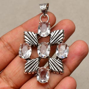 May include: A silver-toned pendant with a cross-shaped design. The pendant features six oval, clear gemstones set within a detailed, ornate frame. The top of the pendant has a decorative bail for a chain. The overall design is intricate and eye-catching.