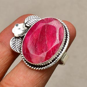 Puede incluir: Un anillo de plata con una gran gema de rubí de corte ovalado. El rubí es de color rojo vibrante y está engastado en un engaste de plata con un borde en forma de cuerda. El anillo tiene un diseño floral y de hojas en el lateral.