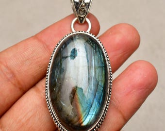 Puede incluir: Colgante ovalado de labradorita con una fianza plateada y un borde decorativo. La piedra preciosa muestra tonos iridiscentes azules, verdes y dorados. El colgante está suspendido de un lazo plateado.