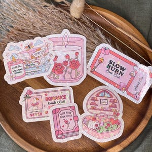 Könnte beinhalten: Eine Sammlung von rosa und irisierenden Aufklebern mit Romantik-Thema. Designs umfassen eine Kerze mit Rosen, ein Buch, eine herzförmige Szene und ein Ticket mit der Aufschrift "Slow Burn Book Club". Andere Aufkleber sagen "County Fair" und "Romance Book Club".