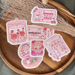 Könnte beinhalten: Eine Sammlung von rosa und schillernden Aufklebern mit Romantik-Thema. Designs umfassen eine Kerze mit Rosen, ein Ticket mit einer Gans und ein Buch mit den Worten "ROMANCE Book Club" und "ROMANTASY BOOK LOVER".