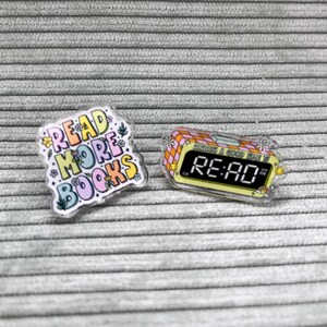 Könnte beinhalten: Zwei bunte Emaille-Pins. Ein Pin zeigt den Text "READ MORE BOOKS" in einer Regenbogen-Schriftart mit Blumen. Der andere Pin ist in Form eines Weckers mit dem Text "RE:AD" auf dem Display.