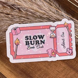 Könnte beinhalten: Ein rosa Aufkleber im Ticketdesign mit dem Text "SLOW BURN Book Club". Der Aufkleber enthält Illustrationen eines Buches, einer Sanduhr, eines Streichholzes und eines herzförmigen Schlosses. Das Design hat einen glitzernden, holografischen Effekt.
