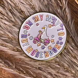 Könnte beinhalten: Runder, weißer Emaille-Pin mit goldenem Rand. Er zeigt eine Comic-Gans auf einem rosa Roller, umgeben von bunten Blumen. Der Text lautet "RUN MY WAY TO BUY MORE BOOKS".
