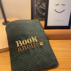 Könnte beinhalten: Eine grüne Cord-Buchhülle mit dem Text "Book aholic", der in goldfarbener Schrift gestickt ist.