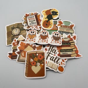 May include: A selection of autumn-themed stickers with book, pumpkin, and leaf designs. Some stickers have text that reads "Cozy with your Kindle", "Books are my happy", and "Her Fall". The stickers are in shades of brown, orange, and cream.
