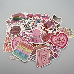 Op de afbeelding: Een verzameling kleurrijke stickers met een leesthema. De stickers tonen boeken, harten en zinnen als "Reading helps your mind bloom" en "Book Girlie". Een roze dinosaurus die een boek leest, is ook afgebeeld.