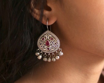May include: Silver dangle earrings with a teardrop-shaped design featuring a peacock motif and red gemstone accents. The earrings have a series of small silver bells hanging from the bottom.