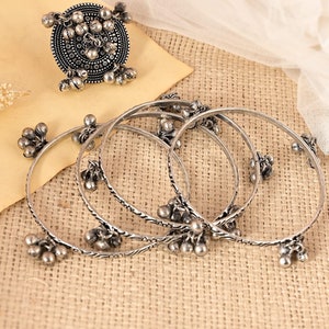 Pode incluir: Conjunto de cinco pulseiras de prata com detalhes decorativos e pequenos sinos pendurados em cada pulseira. As pulseiras est&atilde;o empilhadas sobre uma superf&iacute;cie marrom texturizada.