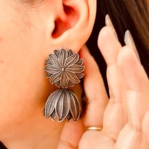 May include: Silver-toned, two-part floral earrings. The top portion is a flat, circular flower design, while the bottom is a bell-shaped flower. The earrings are worn on an ear, with a hand in the frame. The earrings have a vintage or antique aesthetic.