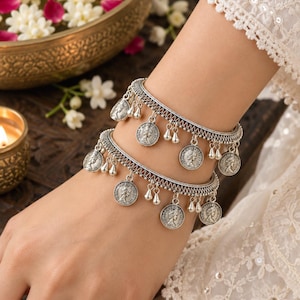 Può includere: Due bracciali rigidi in metallo color argento con un design a fascia testurizzata. Ogni bracciale è decorato con diversi ciondoli circolari a forma di moneta e piccoli pendenti a goccia che pendono uniformemente lungo il bordo inferiore.