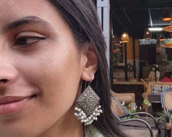May include: Silver-toned, diamond-shaped earrings with intricate filigree patterns and dangling white beads. The earrings are a dangle style, with a hook closure. The design is ornate and detailed, suitable for a bohemian or ethnic aesthetic.