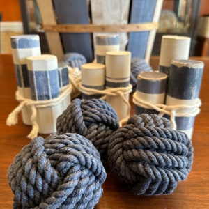 May include: Four dark gray rope balls with a knot design. The balls are sitting on a wooden surface. There are several blue and white striped wooden blocks in the background.