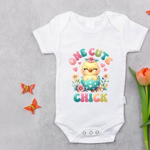 May include: White baby onesie with the text "ONE CUTE CHICK" and a cartoon chick design. The design features a yellow chick hatching from a blue egg, surrounded by flowers and hearts. Decorative orange butterflies and tulips are also present.