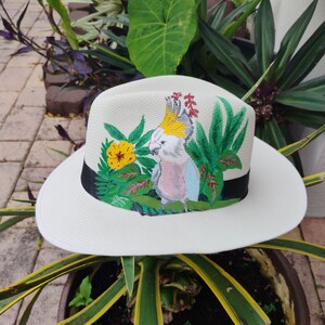 Puede incluir: Un sombrero de paja blanco con un diseño pintado de un cacatúa, plantas tropicales y flores. El pájaro es blanco con una cresta amarilla y detalles rosas. Las plantas son verdes y las flores son amarillas.