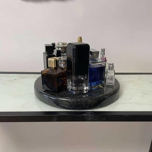 Puede incluir: Una bandeja giratoria de mármol negro con una colección de botellas de vidrio de perfume y colonia. Las botellas son de varias formas y tamaños y tienen detalles dorados.