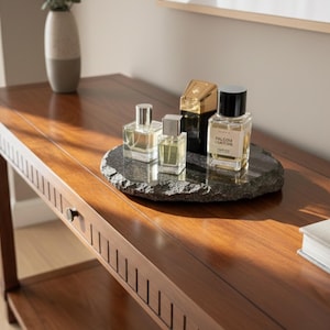 Op de afbeelding: Een houten console met een donkere afwerking toont verschillende parfumflesjes op een donkere stenen tray. De flesjes hebben verschillende vormen en maten, met helder glas en zilveren of zwarte doppen. Een decoratieve vaas staat op de tafel.