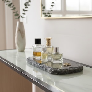 May include: A collection of perfume bottles of varying sizes and shapes, displayed on a dark gray, rectangular stone tray. The bottles are clear, showcasing the amber and light-colored liquids inside. A white vase with green stems is in the background.