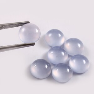 Puede incluir: Una colecci&oacute;n de gemas redondas de color azul p&aacute;lido. Una gema es sostenida por unas pinzas plateadas, mientras que otras seis est&aacute;n dispersas cerca. Las gemas tienen una superficie lisa y pulida y una apariencia transl&uacute;cida, con un efecto suave y brillante. Las gemas est&aacute;n sobre un fondo blanco.