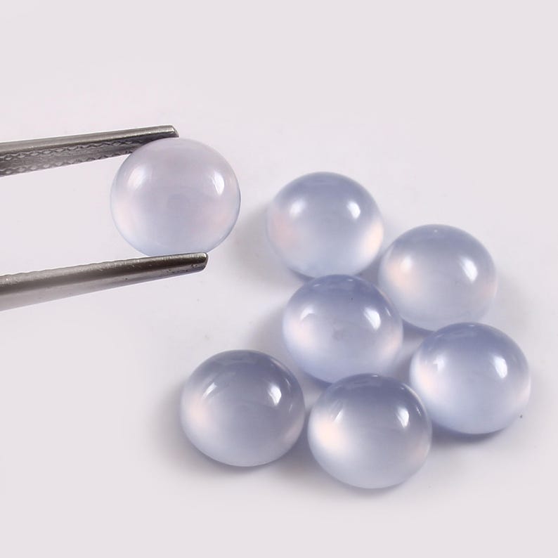 Puede incluir: Una colecci&oacute;n de gemas redondas de color azul p&aacute;lido. Una gema es sostenida por unas pinzas plateadas, mientras que otras seis est&aacute;n dispersas cerca. Las gemas tienen una superficie lisa y pulida y una apariencia transl&uacute;cida, con un efecto suave y brillante. Las gemas est&aacute;n sobre un fondo blanco.