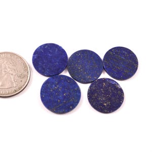 Op de afbeelding: Vijf ronde, gepolijste lapis lazuli stenen in verschillende blauwtinten, met gouden vlekken. Een munt ligt naast de stenen ter vergelijking van de grootte. De stenen liggen op een wit oppervlak. De stenen hebben een diameter van ongeveer 2,5 cm.