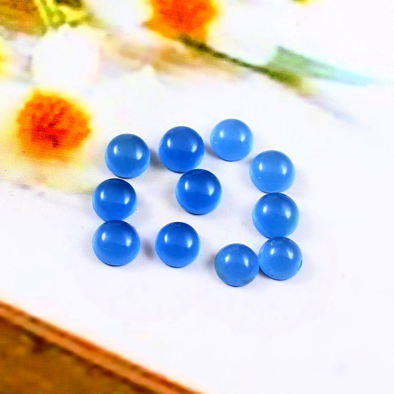 Może przedstawiać: Kolekcja jedenastu okrągłych, wypolerowanych, błękitnych kamieni szlachetnych. Kamienie szlachetne ułożone są w kształt kwadratu, z kilkoma rozrzuconymi na krawędziach. Kamienie mają gładką, błyszczącą powierzchnię.