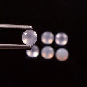 May include: A close-up of several round, pale gemstones. One is held by tweezers, showcasing its faceted surface. The other gemstones are scattered, with a soft, translucent appearance. The background is a solid black, highlighting the gems.