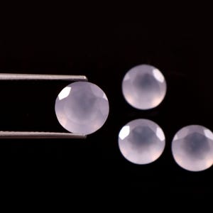 May include: Four pale lavender, faceted, round gemstones against a black background. One gem is held by tweezers. The gems have a polished, reflective surface, and are likely used for jewelry making.