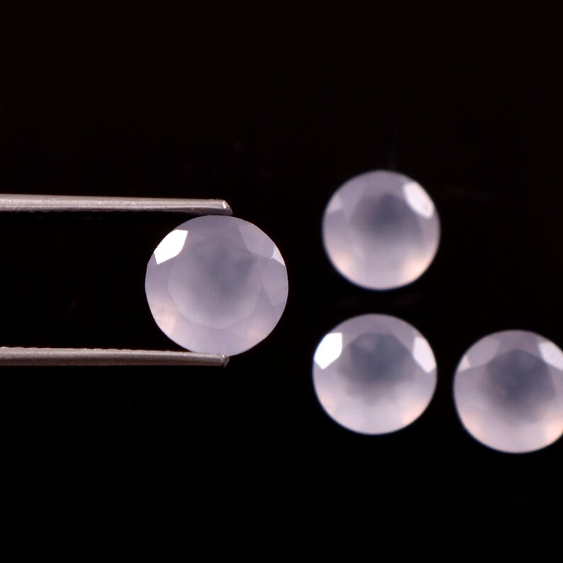 May include: Four pale lavender, faceted, round gemstones against a black background. One gem is held by tweezers. The gems have a polished, reflective surface, and are likely used for jewelry making.